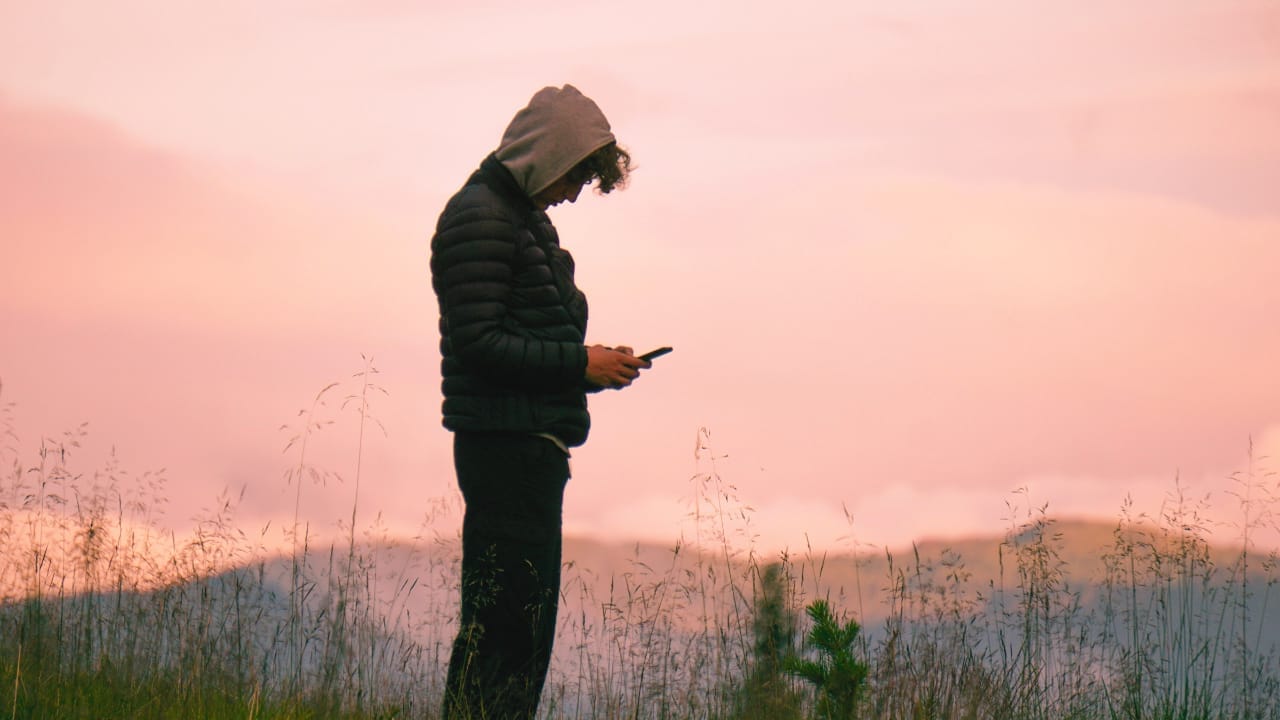 A young person checks their phone outdoors, surrounded by plants, mountains in the background and clouds