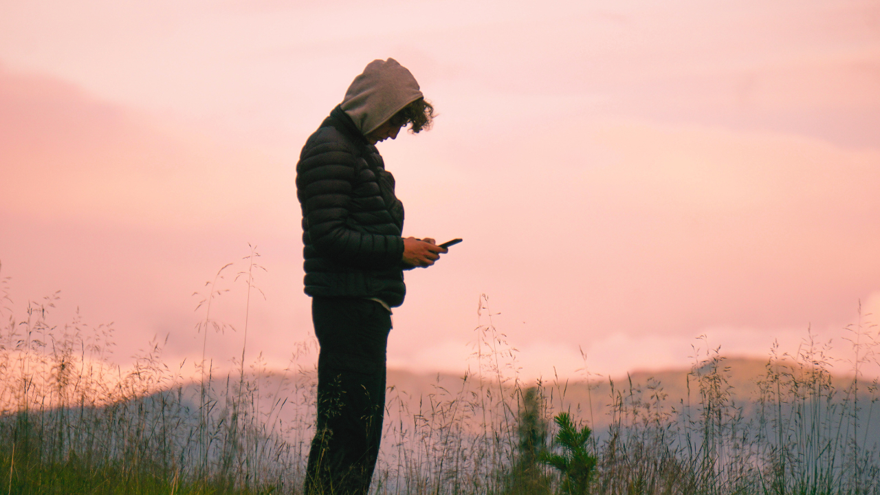 A young person checks their phone outdoors, surrounded by plants, mountains in the background and clouds